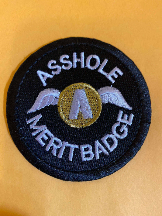 Black round merit badge patch with white embroidered text "Asshole Merit Badge" and a gold winged emblem in the center