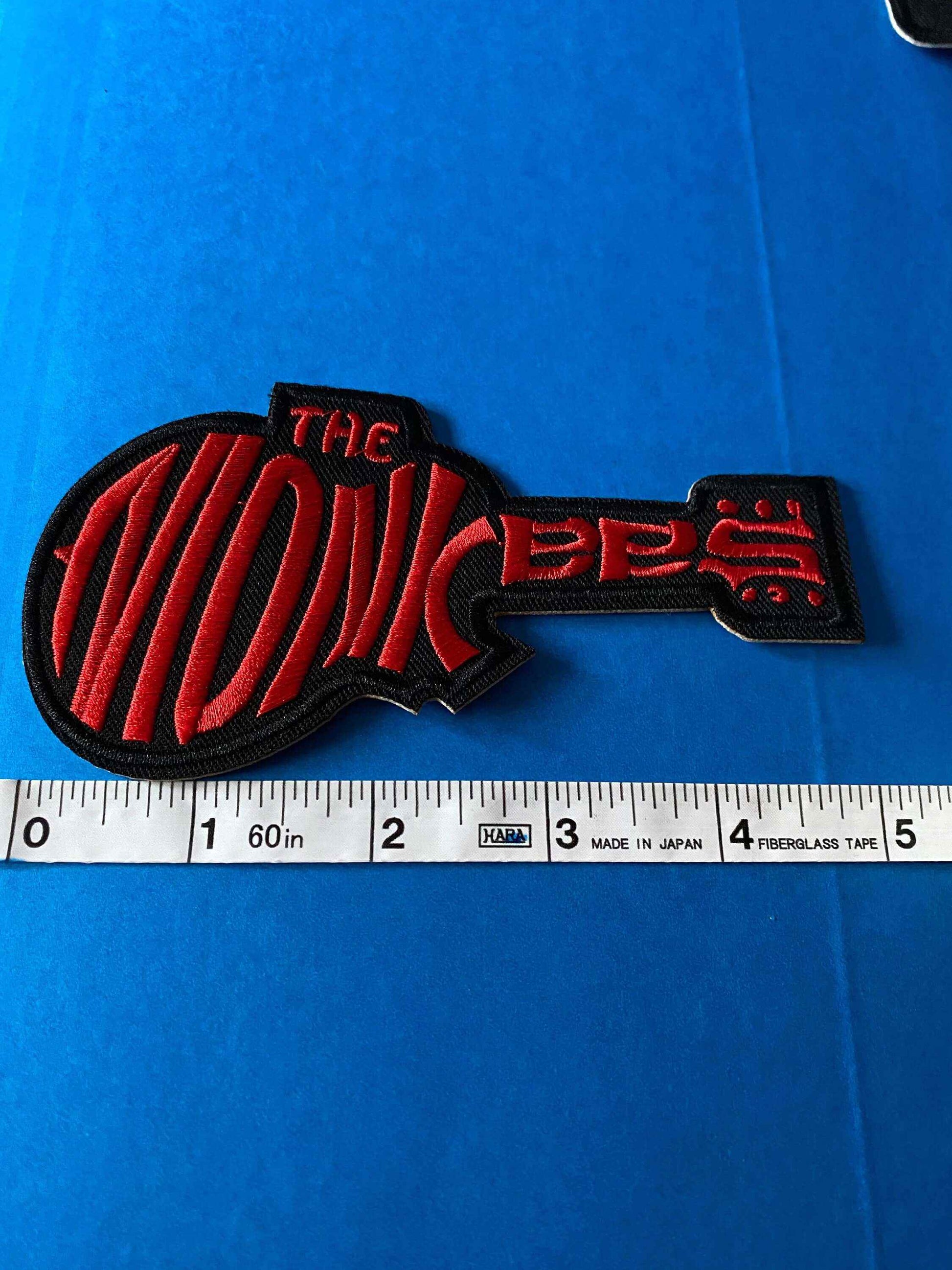 The Monkees logo patch embroidered in red and black shaped like a guitar with a ruler for size reference on a blue background