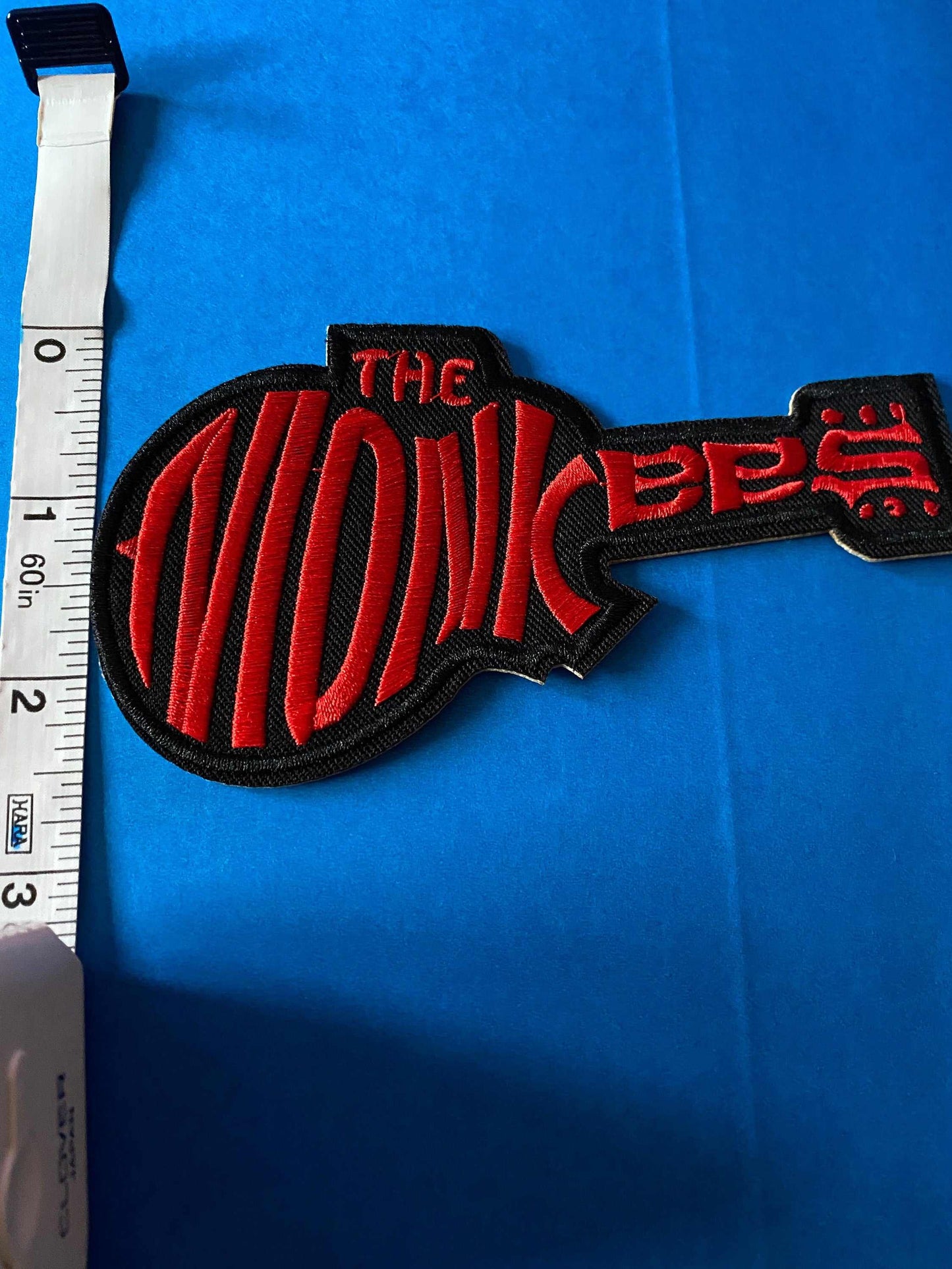 Red and black embroidered Monkees logo patch shaped like a guitar next to a measuring tape on blue background