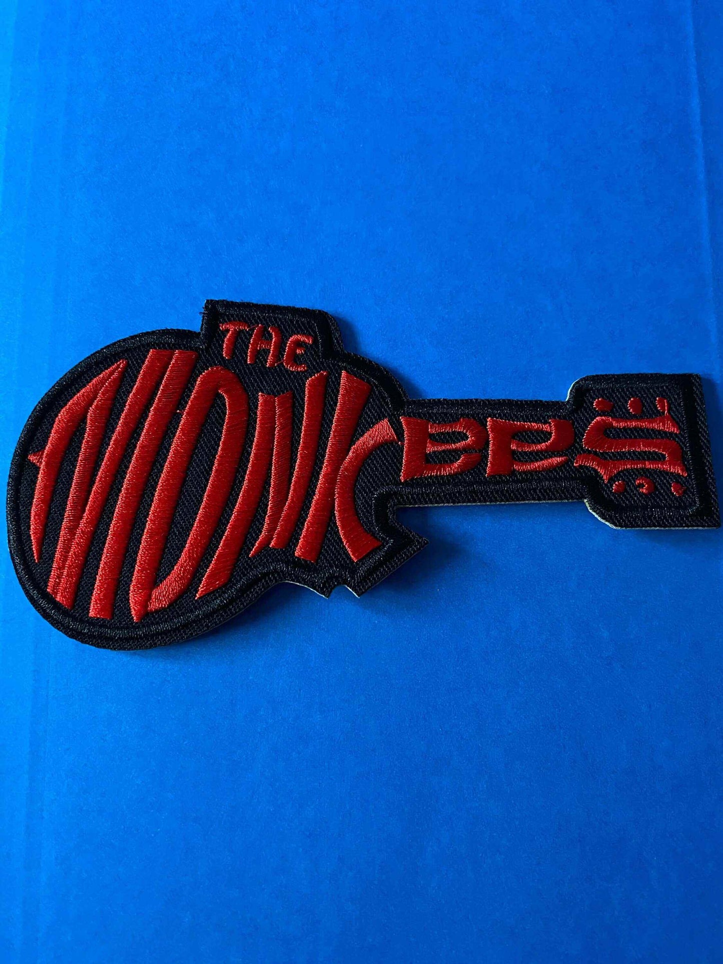 The Monkees logo patch in red and black embroidered design shaped like a guitar on a blue background